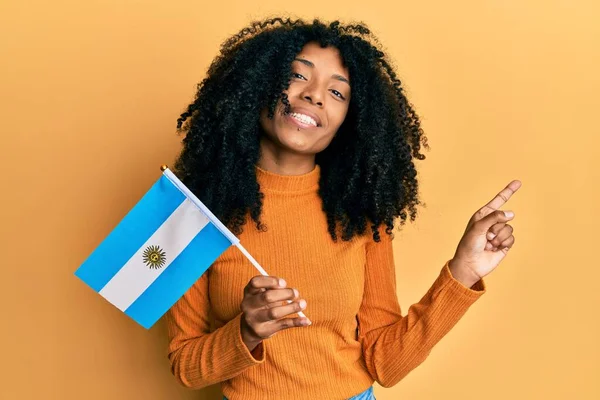 African american woman with afro hair holding argentina flag smiling happy pointing with hand and finger to the side