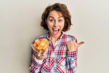 Patates cipsi tutan genç esmer kadın başparmağıyla yan tarafı işaret ediyor ve ağzı açık gülümsüyor. 