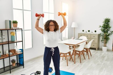 Orta yaşlı İspanyol kadın gülümsüyor. Eğitimine özgüveni tam. Evde dambıl kullanıyor.
