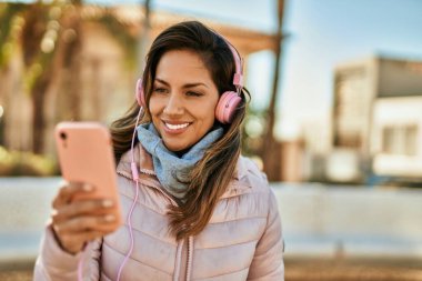 Genç İspanyol kadın şehirde akıllı telefon ve kulaklık kullanarak mutlu bir şekilde gülümsüyor..