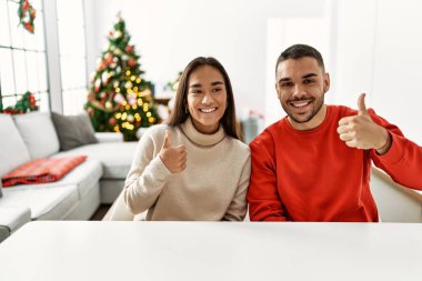 Genç İspanyol çift Noel ağacının yanında oturmuş gülümsüyor, mutlu ve pozitif, başparmağıyla mükemmel ve onay işareti yapıyor. 