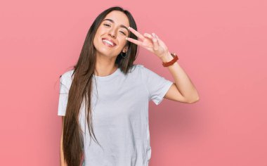 Gündelik beyaz tişört giyen genç İspanyol kız barış sembolü yapıyor. Yüzünde parmaklar, gülümseyerek zaferi gösteriyor. 