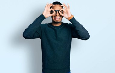Genç Afro-Amerikan adam günlük kıyafetler ve gözlük takıyor dürbün gibi jest yapıyor, dili dışarı çıkarıyor, gözleri parmaklarından bakıyor. çılgın ifade. 