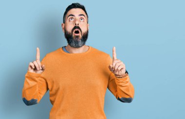 Kış süveteri giyen İspanyol sakallı adam şaşkın şaşkın yukarı baktı ve parmaklarıyla işaret ederek kollarını kaldırdı.. 