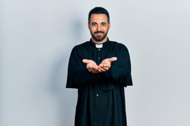 Sakallı, Katolik rahip cübbesi giymiş, elleri avuçlarında gülümseyen yakışıklı İspanyol bir adam el hareketi yapıyor. Bekleme ve koruma 
