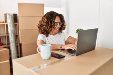 Orta yaşlı İspanyol kadın yeni evinde dizüstü bilgisayar kullanarak gülümsüyor.