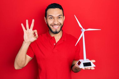 Sakallı İspanyol adam yenilenebilir elektrik için güneş değirmeni tutuyor. Parmaklarıyla imza atıyor, gülümseyerek mükemmel bir sembol oluşturuyor. 