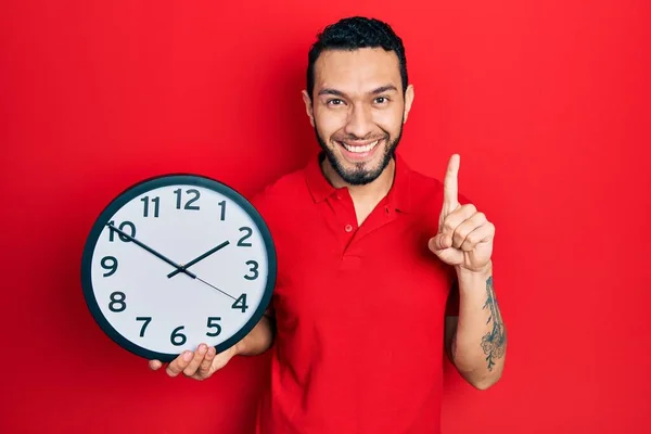 Latynos Brodą Trzymający Wielki Zegar Uśmiechający Się Pomysłem Lub Pytaniem — Zdjęcie stockowe