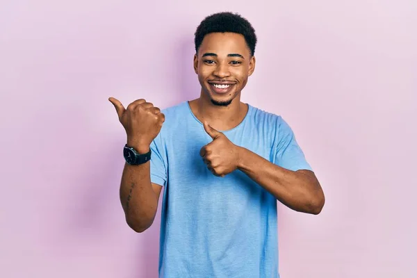 手と親指を上げて後ろを指差すカジュアルブルーのTシャツを着た若いアフリカ系アメリカ人の男は 自信を持って笑って — ストック写真