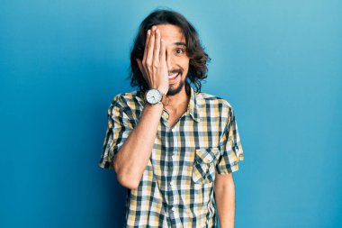 Latin asıllı genç adam gündelik giysiler giyiyor. Bir gözünü eliyle kapatıyor. Yüzünde kendine güvenen bir gülümseme ve sürpriz bir duygu var.. 