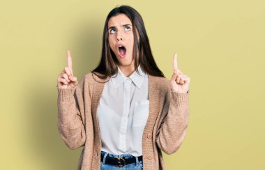 Genç esmer, günlük beyaz gömlek ve ceket giyen genç şaşkın ve şaşırmış yukarı bakıyor ve parmaklarıyla işaret ediyor ve kollarını kaldırıyor.. 