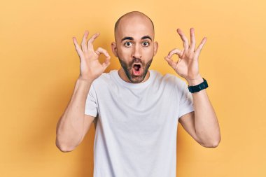 Gündelik beyaz tişört giyen genç kel adam şaşırmış ve şok olmuş bir şekilde parmaklarıyla onay sembolü yapıyor. çılgın ifade 