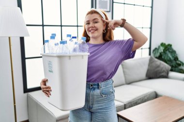 Geri dönüşüm sepetini elinde tutan genç, kızıl saçlı bir kadın. Elinde plastik şişeler var. Tek parmağıyla kafasına gülümsüyor. 