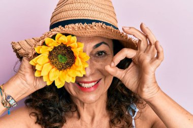 Orta yaşlı İspanyol kadın, yaz şapkası takıyor, elinde ayçiçeği var. Parmaklarıyla işaret veriyor, gülümsüyor, mükemmel bir sembol oluşturuyor. 