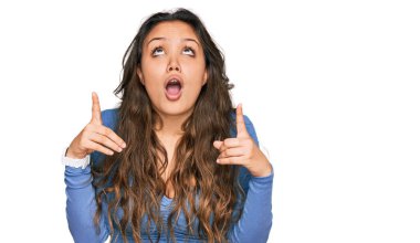 Gündelik kıyafetler giyen genç İspanyol kız şaşkın şaşkın yukarı bakıyor ve parmaklarıyla işaret ediyor ve kollarını kaldırıyor.. 