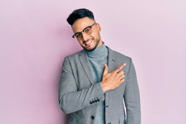 İş kıyafetleri giymiş genç bir Arap. Gülümsüyor, eli ve parmağı yana doğru uzanıyor. 