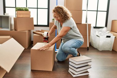 Orta yaşlı beyaz kadın yeni evinde karton kutu paketlerken mutlu gülümsüyor..