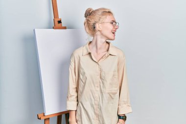 Güzel, beyaz, sarışın bir kadın boyacı sehpasının yanında duruyor yüzünde gülümsemeyle, doğal bir ifadeyle yan yana bakıyor. Kendinden emin gülen. 