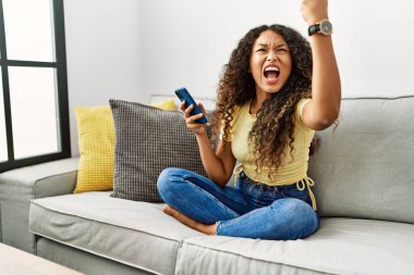 Güzel İspanyol kadın evinde kanepede oturuyor. Akıllı telefon kullanıyor. Kızgın ve kızgın. Öfkeyle bağırırken yumruğunu kaldırıyor. öfke ve agresif konsept. 