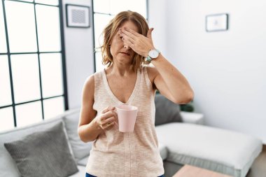 Orta yaşlı bir kadın evde kahve içiyor. Elleriyle gözlerini kapatıyor. Ciddi ve üzgün görünüyor. Görme, saklama ve reddetme konsepti 