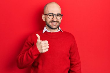 Sıradan kıyafetler ve gözlük takan genç kel bir adam mutlu bir el hareketi yapıyor. Kameraya bakıp başarıyı gösteren ifade. 