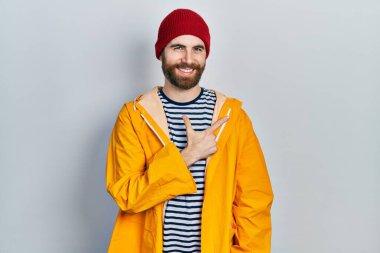 Sakallı, sarı yağmurluk giyen, yüzünde bir gülümseme olan beyaz bir adam, mutlu ve doğal bir ifadeyle elini ve parmağını yana kaldırdı. 