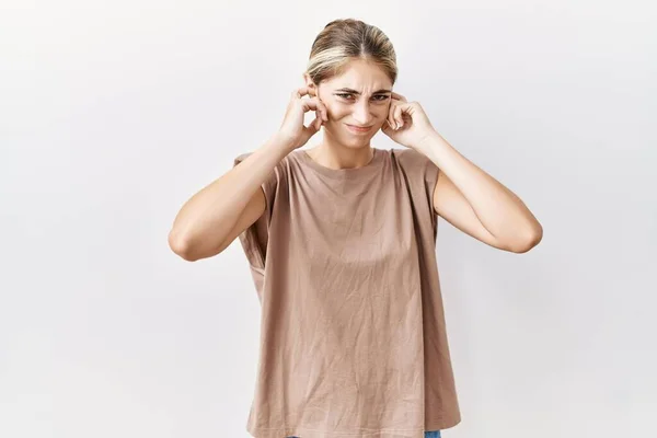 孤立した背景の上に立つ若いブロンドの女性は 大きな音楽のノイズのためにいらいらする表情で指で耳を覆う 聴覚障害の概念 — ストック写真