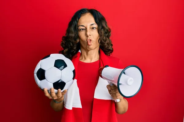 Middle age hispanic woman football hooligan holding ball and using megaphone making fish face with mouth and squinting eyes, crazy and comical.