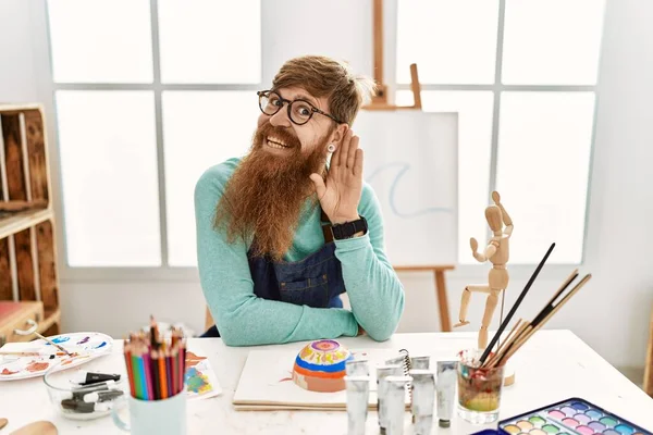 红头发的男人 留着长胡子 在艺术工作室里画着黏土碗 面带微笑 侧耳倾听谣言或流言蜚语 聋的概念 — 图库照片