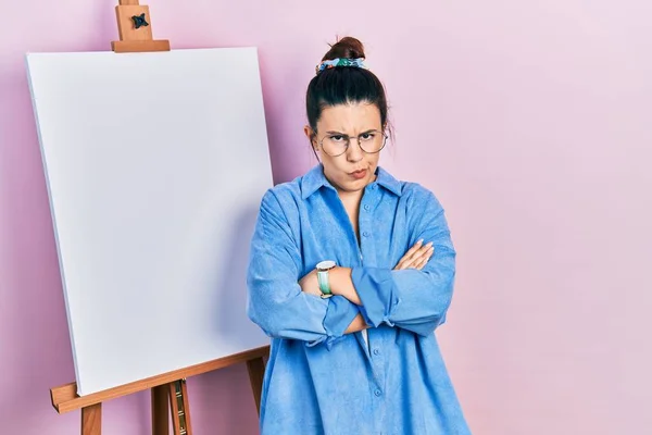 画家のイーゼルのそばに立つ若いヒスパニック系の女性は 両腕を組んで顔に懐疑的で緊張し 不承認の表現に立っています 否定的な人 — ストック写真