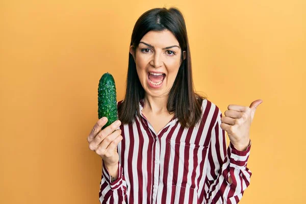 Joven Mujer Hispana Sosteniendo Pepino Apuntando Con Pulgar Hacia Costado —  Fotos de Stock