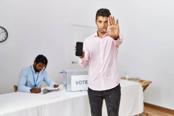 Young hispanic men at political campaign election holding smartphone with open hand doing stop sign with serious and confident expression, defense gesture