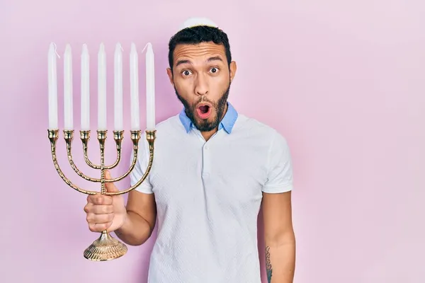 髭を生やしているヒスパニック系の男Menora Hanukkaユダヤ人のキャンドル怖がって驚きのために口を開けて驚いて 不信の顔 — ストック写真