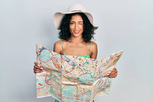 Young latin girl wearing summer hat holding city map winking looking at the camera with sexy expression, cheerful and happy face.
