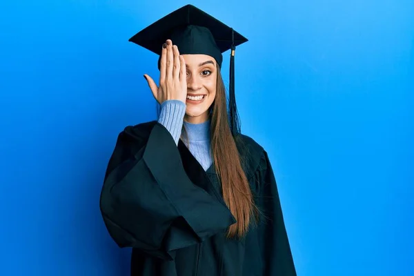卒業キャップと式のローブを身に着けている美しいブルネットの若い女性が手で一つの目を覆い 顔に自信を持って笑顔と驚きの感情 — ストック写真