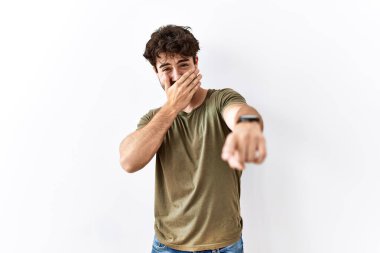 İzole edilmiş beyaz arka planda duran İspanyol adam sana gülüyor, eli ağzında kamerayı işaret ediyor, utanç ifadesi var. 