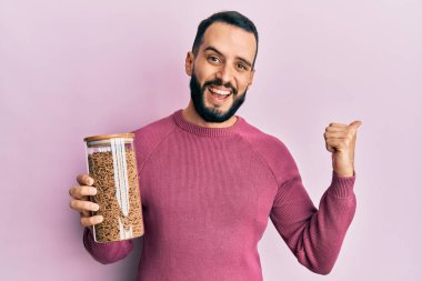 Sakallı, sağlıklı tahıl gevreği kavanozu tutan genç bir adam başparmağını yana uzatıyor ve ağzı açık gülümsüyor. 