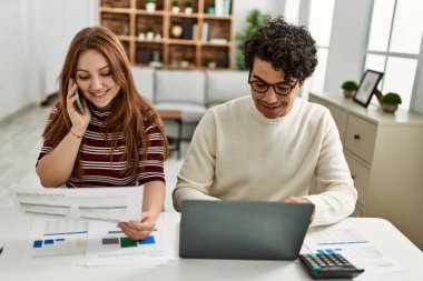 Genç çift dizüstü bilgisayar kullanıyor ve akıllı telefonla konuşuyor. Evde masanın üzerinde oturuyor..