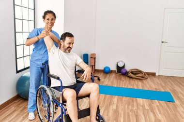 Orta yaşlı bir adam ve kadın, fizik tedavi kliniğinde tekerlekli sandalyeye bağlı olarak rehabilitasyon seansı yapıyorlar.