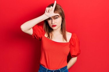 Kırmızı tişörtlü kızıl saçlı genç bir kadın. Alnında parmakları olan insanlarla dalga geçiyor. Ezik jestler yapıyor.. 