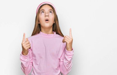 Güzel esmer, balıkçı yaka kazak giyen küçük kız şaşırmış ve şaşırmış bir şekilde yukarı bakıyor ve parmaklarıyla işaret ediyor ve kollarını kaldırıyor.. 