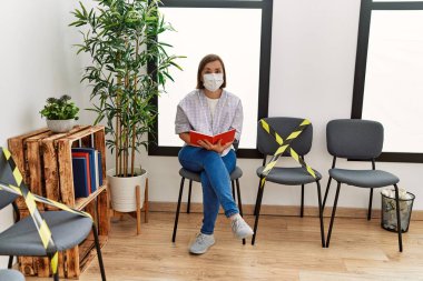 Orta yaşlı İspanyol kadın güvenlik maskesi takıp bekleme odasında kitap okuyor.