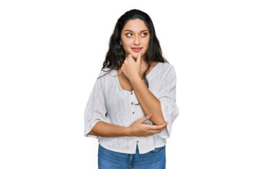 Elleri çenesinde gündelik kıyafetler giyen esmer genç bir kadın soru sormayı düşünüyor, düşünceli bir ifade. Düşünceli bir yüzle gülümsüyor. şüphe kavramı. 