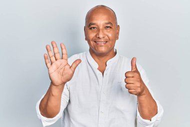 Orta yaşlı, gündelik kıyafetler giyen Latin bir adam altı numarayı gösterip işaret ederken kendinden emin ve mutlu bir şekilde gülümsüyor.. 