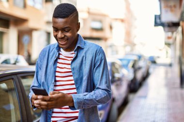 Genç Afrikalı Amerikalı adam şehirde akıllı telefon kullanarak mutlu bir şekilde gülümsüyor..