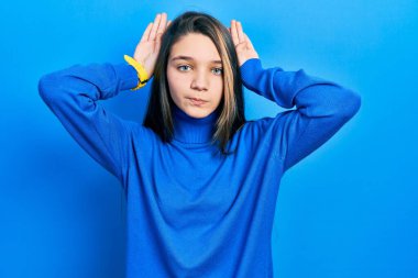 Balıkçı yaka kazak giyen, tavşan kulağı jestleri yapan, elleri avuçlarında kuşkucu ve şüpheci görünen genç esmer kız. Paskalya tavşanı konsepti. 