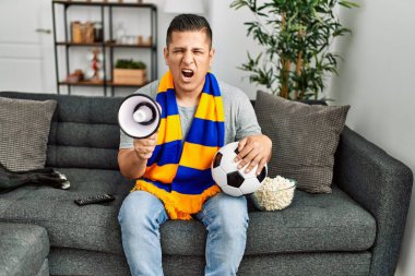 Genç İspanyol holigan adam futbol takımını destekliyor evdeki kanepede oturuyor..