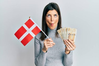 Danimarka bayrağı ve krone banknotları tutan genç İspanyol kadın ağızlarıyla ve şaşı gözleriyle balık yüzü yapıyor, çılgın ve komik.. 