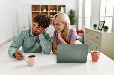 Laptop ve akıllı telefon kullanan genç bir çift evde kahve içiyor..