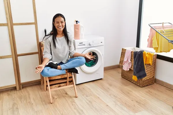 Young latin woman doing yoga waiting for washing machine at laundry room